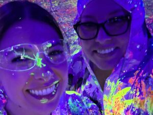 Two people smiling under blacklight, wearing clear safety goggles and protective clothing splattered with neon paint, creating a vibrant, colorful glow in the dark environment.