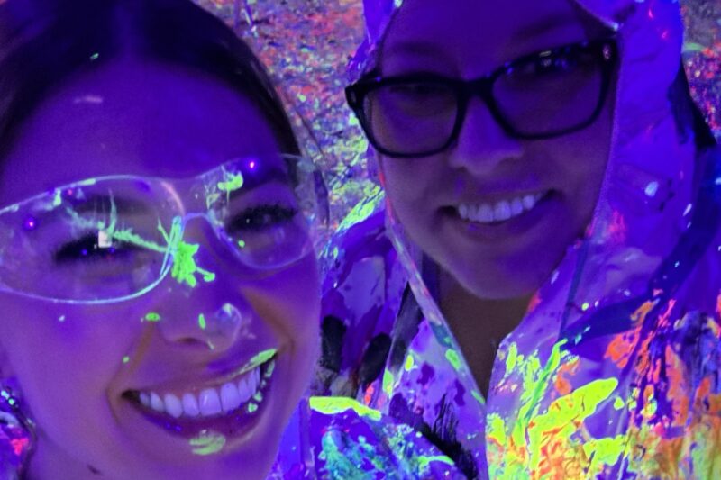 Two people smiling under blacklight, wearing clear safety goggles and protective clothing splattered with neon paint, creating a vibrant, colorful glow in the dark environment.