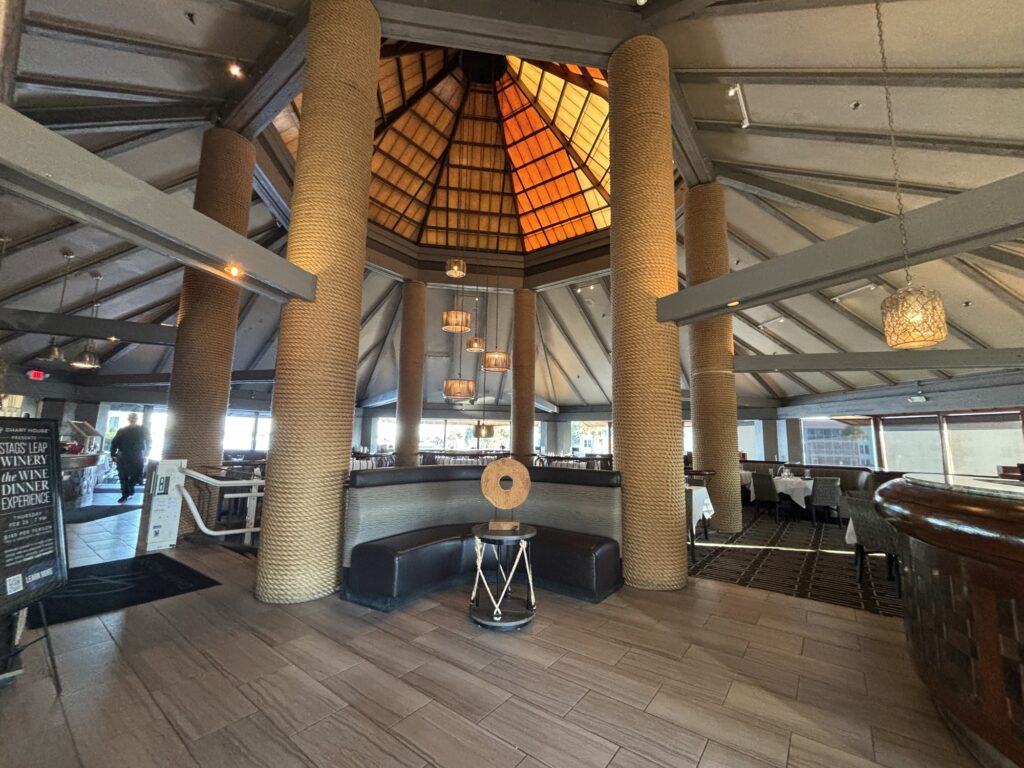 The spacious restaurant interior of the Chart House with high vaulted ceilings, large textured columns, modern hanging lights, and a circular seating area; tables with white cloths are visible in the background.