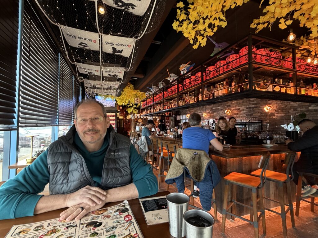 A man sits at a table with a menu in a cozy bar with brick walls, warm lights, hanging decor, and people seated at the counter. Yellow leaves and Japanese banners hang from the ceiling.