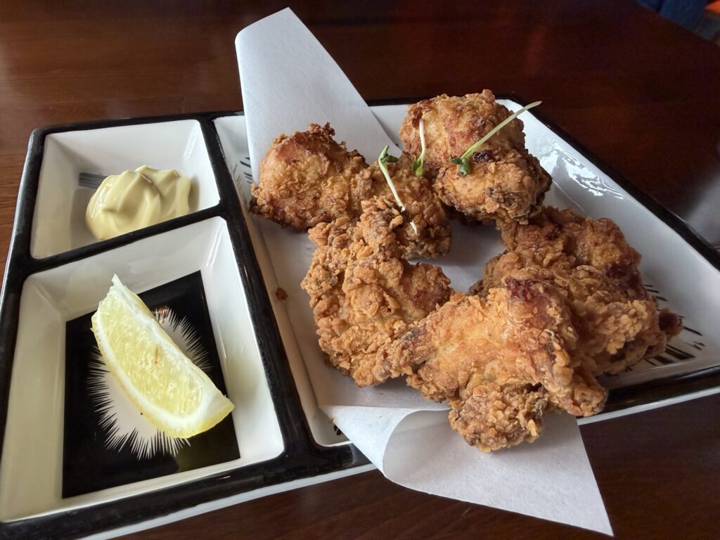 Four pieces of golden-brown fried chicken are served on a tray with a wedge of lemon and a side of creamy dipping sauce, presented on white paper and ceramic dishes on a wooden table.