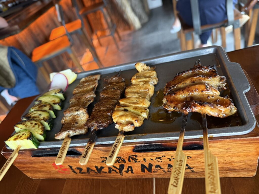 A wooden tray holds six skewers of grilled food, including vegetables, meats, and glazed chicken wings, displayed on a metal serving plate at a restaurant with orange chairs visible in the background.