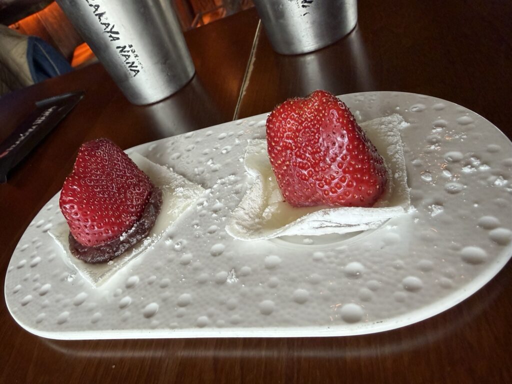 Two pieces of mochi topped with large strawberries are served on a white plate with a textured pattern. Cups and a menu are visible in the background on a dark wooden table.