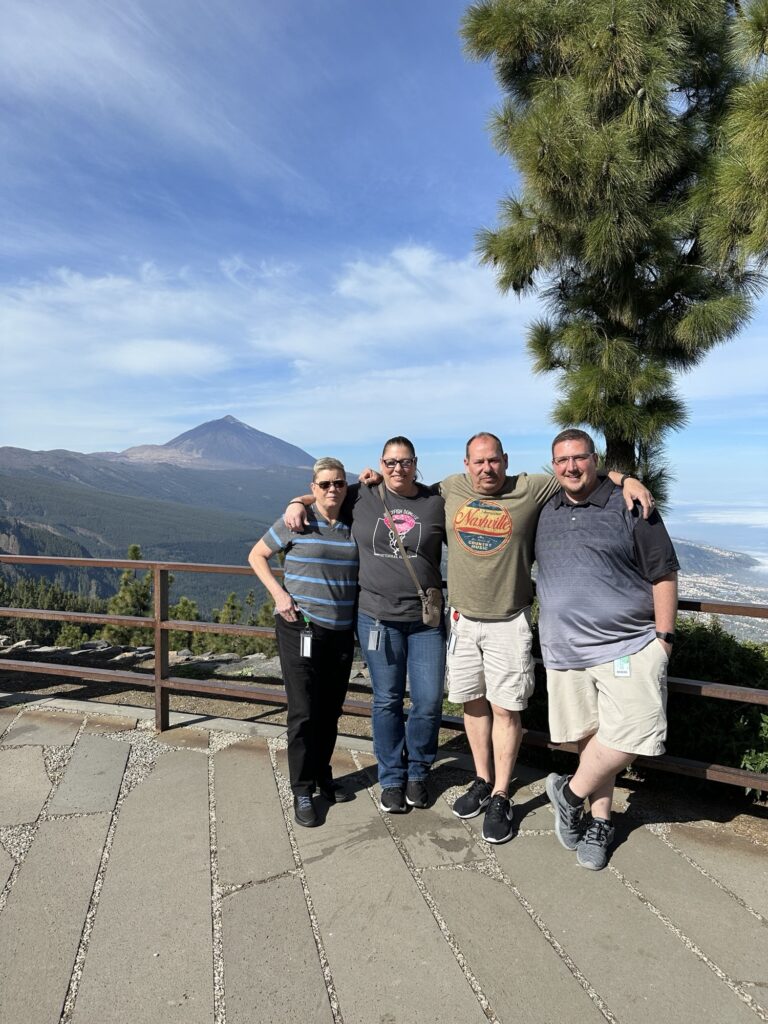 Four people stand side by side, smiling at the camera on a terrace with mountains, trees, and a blue sky in the background. They are dressed casually and the setting appears sunny and scenic.
