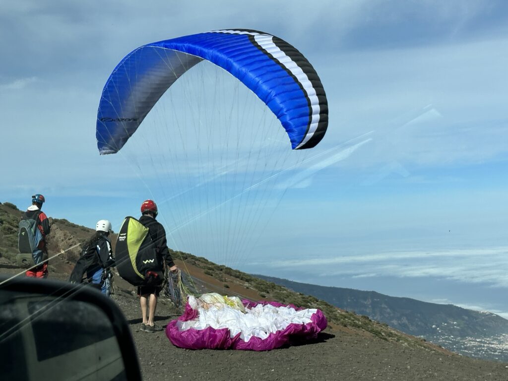 Several people prepare for paragliding on a hillside, with one parachute laid out on the ground and another blue parachute catching wind. The sky is partly cloudy, and mountains are visible in the distance. A car window frames the foreground.