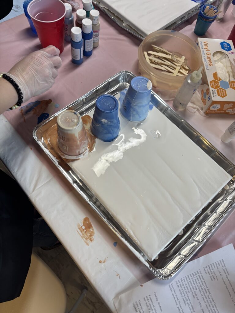A person wearing a glove prepares for a paint-pouring art project, with three paint-filled cups placed upside down on a white canvas in a tray. Bottles of paint, gloves, and mixing sticks are nearby on a pink-covered table.