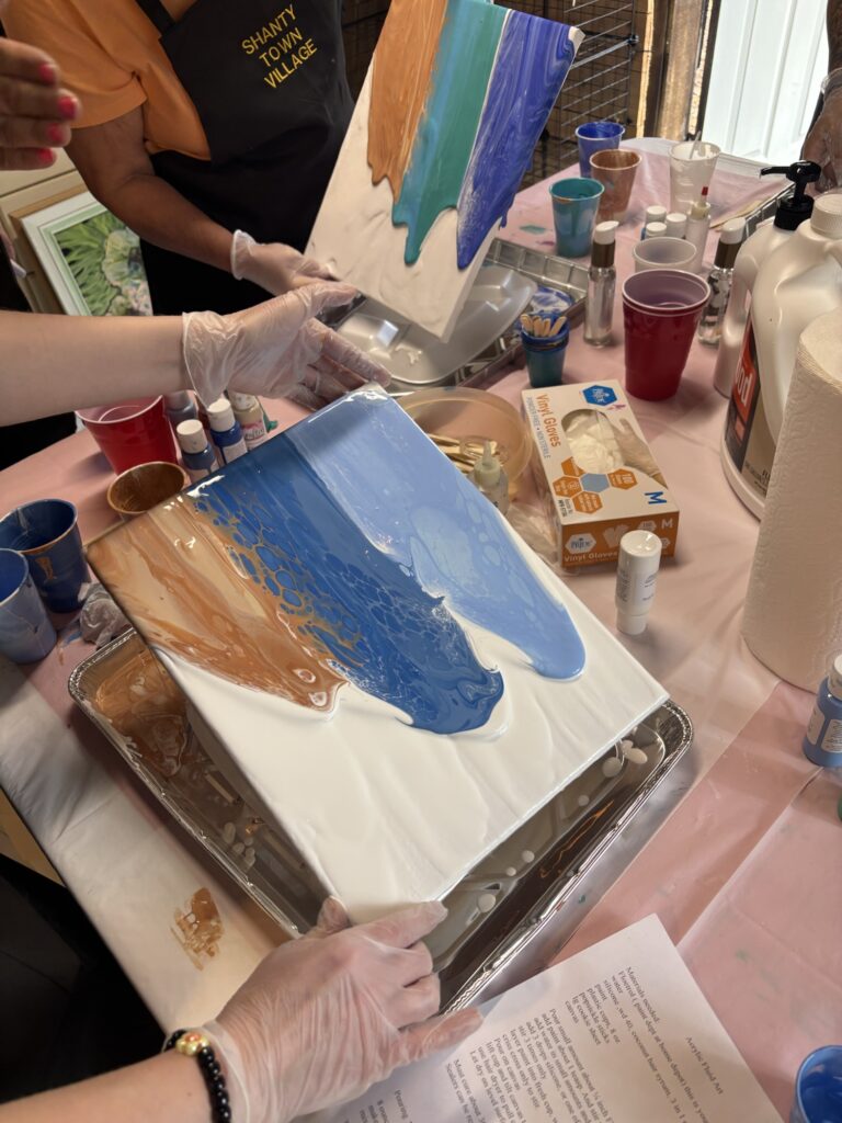 Two people in gloves hold a canvas covered in blue, white, and brown paint, creating a poured paint art piece. Art supplies, cups, and another painted canvas are on the table. A paint instruction sheet is visible.