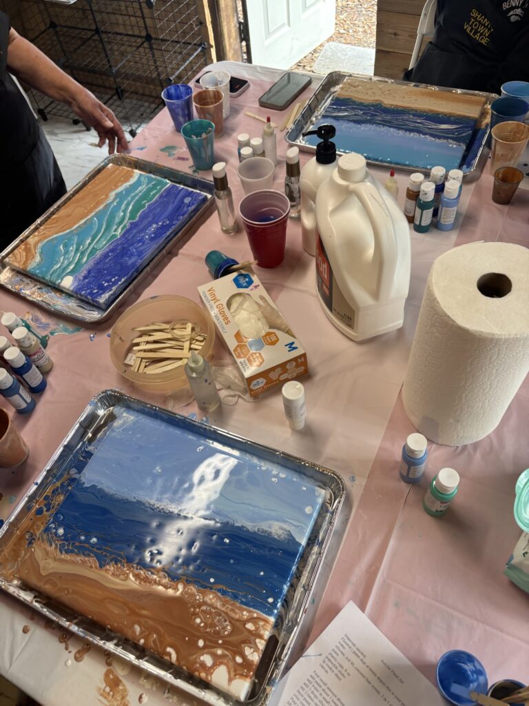 Three trays with fluid acrylic pour paintings resembling ocean waves sit on a pink-covered table, surrounded by paints, cups, stir sticks, gloves, a paper towel roll, and a large bottle of white fluid. Multiple people are painting.