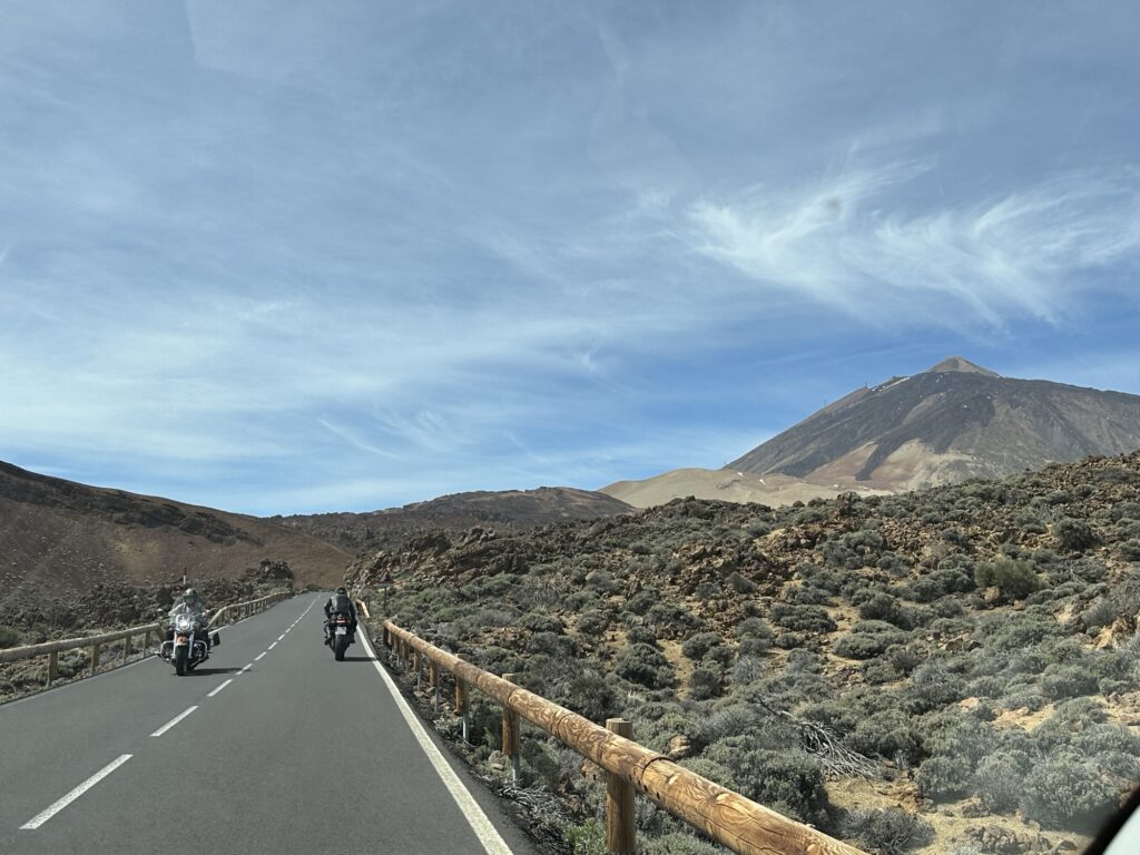Two motorcyclists ride on a paved road through a rocky, mountainous landscape under a blue sky with wispy clouds; a large mountain rises in the background.