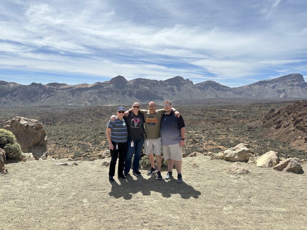 Four people stand close together, smiling, with arms around each other in front of a rocky, mountainous landscape under a blue sky with wispy clouds. The terrain is dry and rugged, with distant peaks in the background.