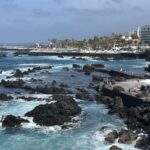 Rocky coastline with turquoise waves crashing against dark volcanic rocks, a crowded boardwalk with people, and a seaside town with palm trees and white buildings in the background under a partly cloudy sky.