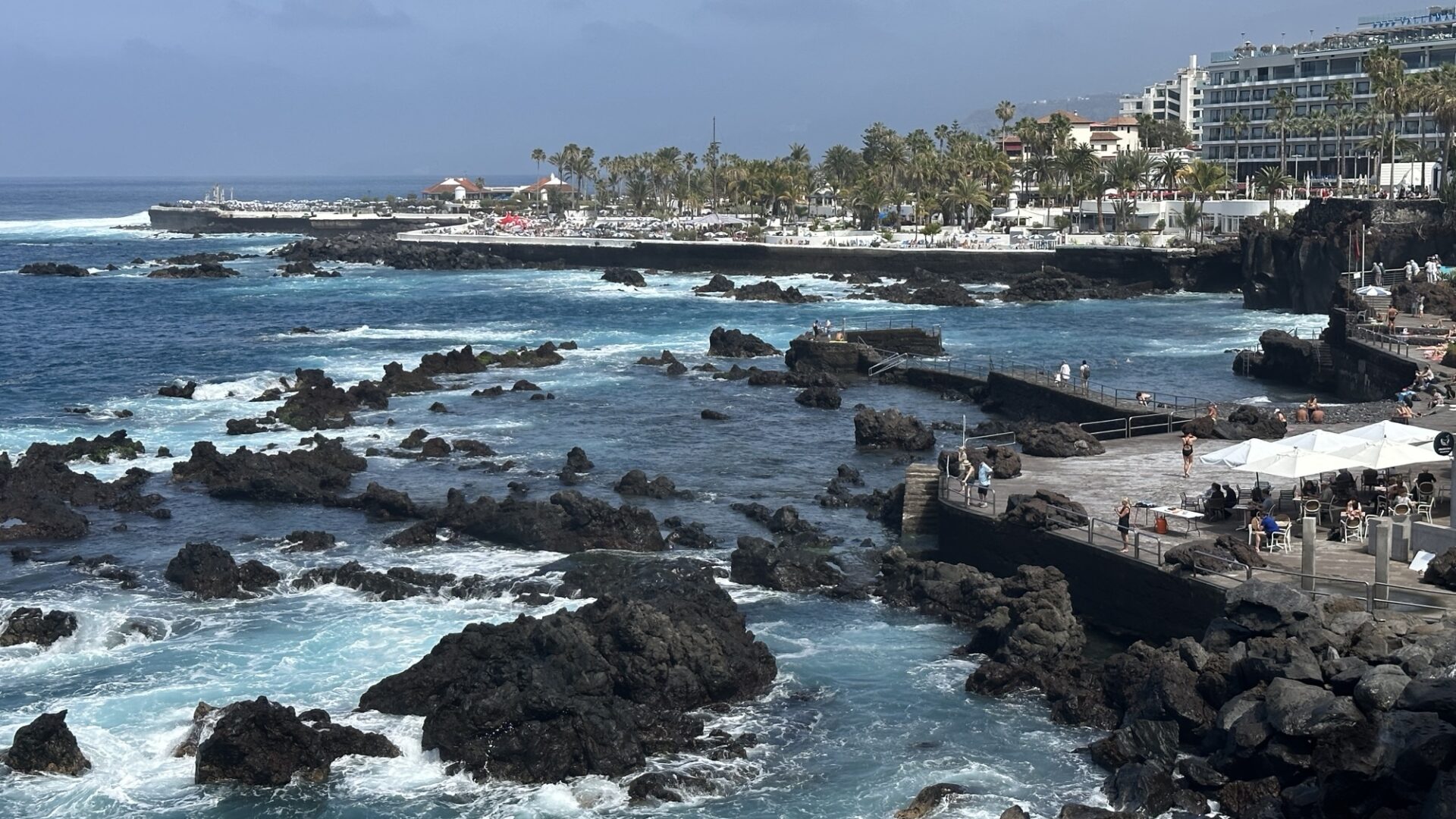 Rocky coastline with turquoise waves crashing against dark volcanic rocks, a crowded boardwalk with people, and a seaside town with palm trees and white buildings in the background under a partly cloudy sky.