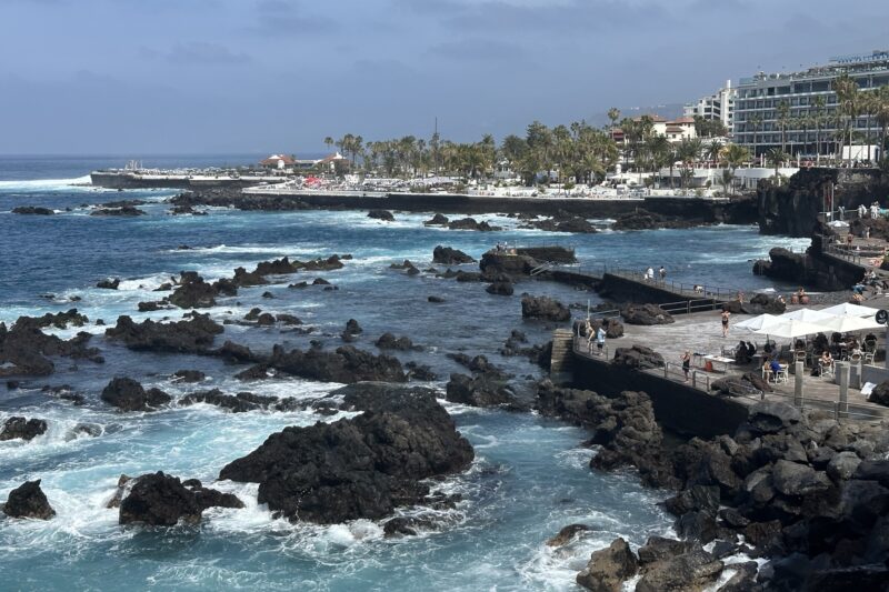 Rocky coastline with turquoise waves crashing against dark volcanic rocks, a crowded boardwalk with people, and a seaside town with palm trees and white buildings in the background under a partly cloudy sky.