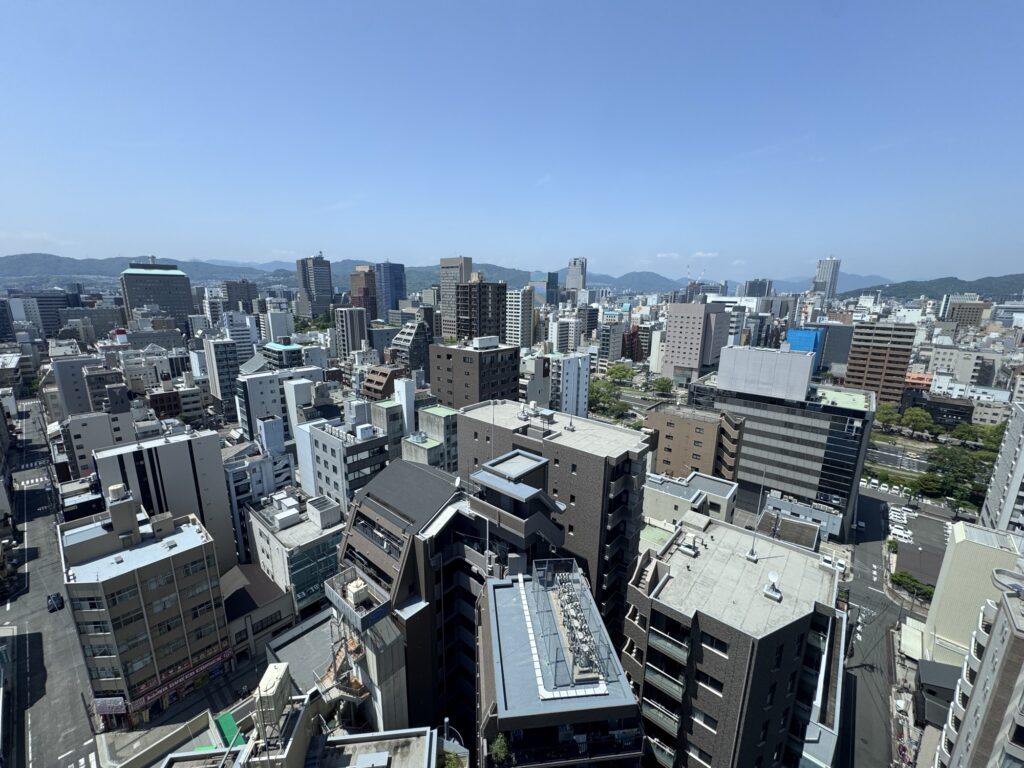 A wide-angle view of a cityscape with numerous modern buildings and apartments under a clear blue sky, mountains visible in the distance, and sunlight casting shadows across the scene.