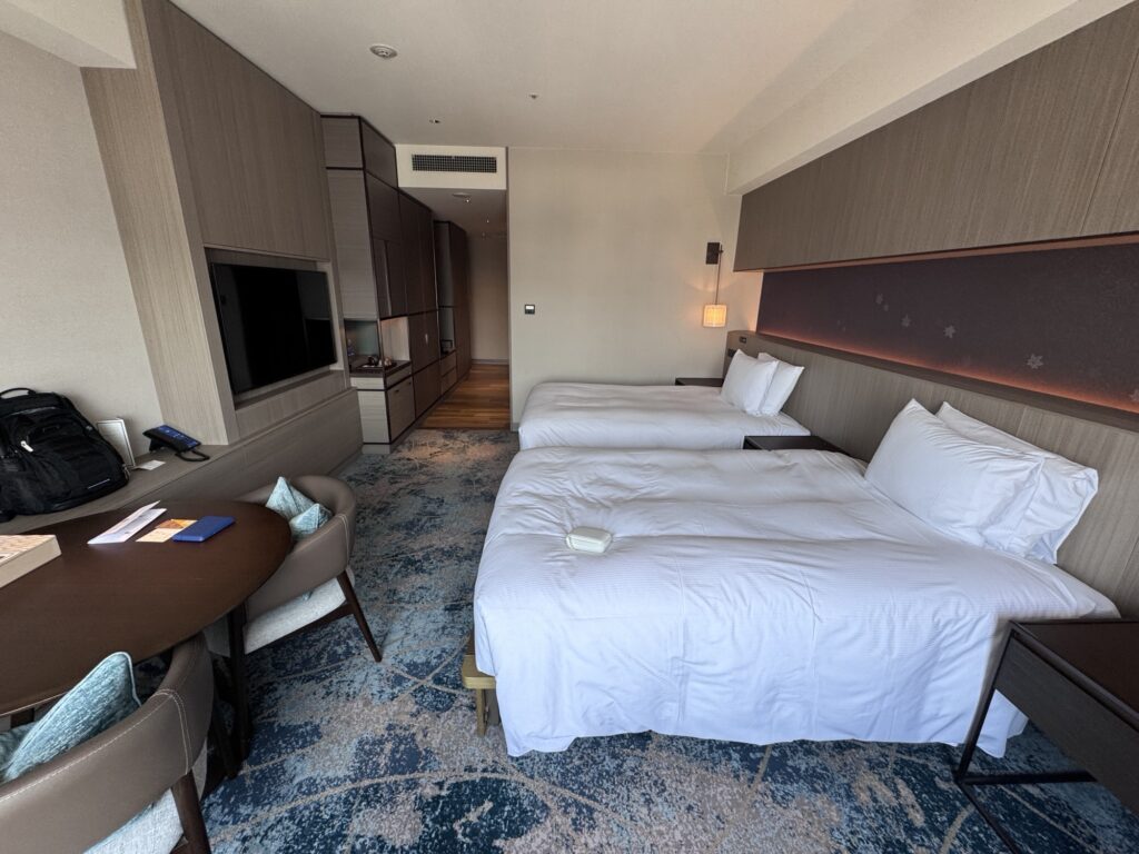 Modern hotel room with two neatly made single beds, a blue patterned carpet, a work desk with chairs, a TV on the wall, a small sitting area, and soft natural light coming in from a window out of view.