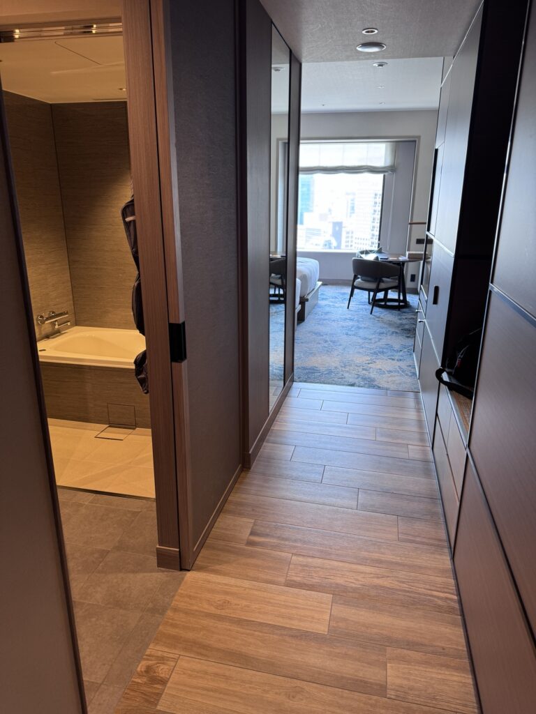 A hotel room hallway with wood floors leads to a modern bedroom with a large window. On the left, an open door reveals a bathroom with a bathtub. Natural light brightens the space.