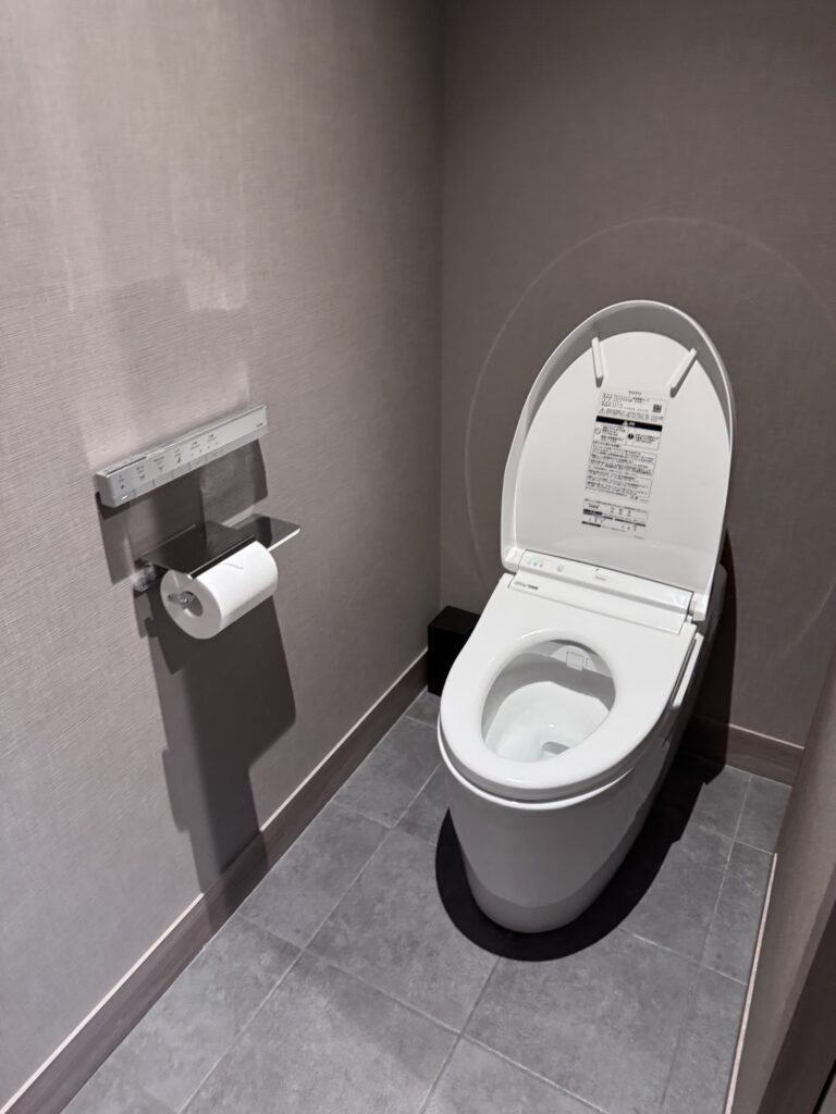 A modern, high-tech toilet with an open lid and control panel on the side, next to a wall-mounted toilet paper holder, in a small, tiled restroom.