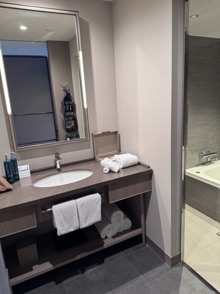 A modern hotel bathroom with a sink and mirror, neatly folded towels on the counter and shelves, toiletries by the sink, and a bathtub visible through a glass door.