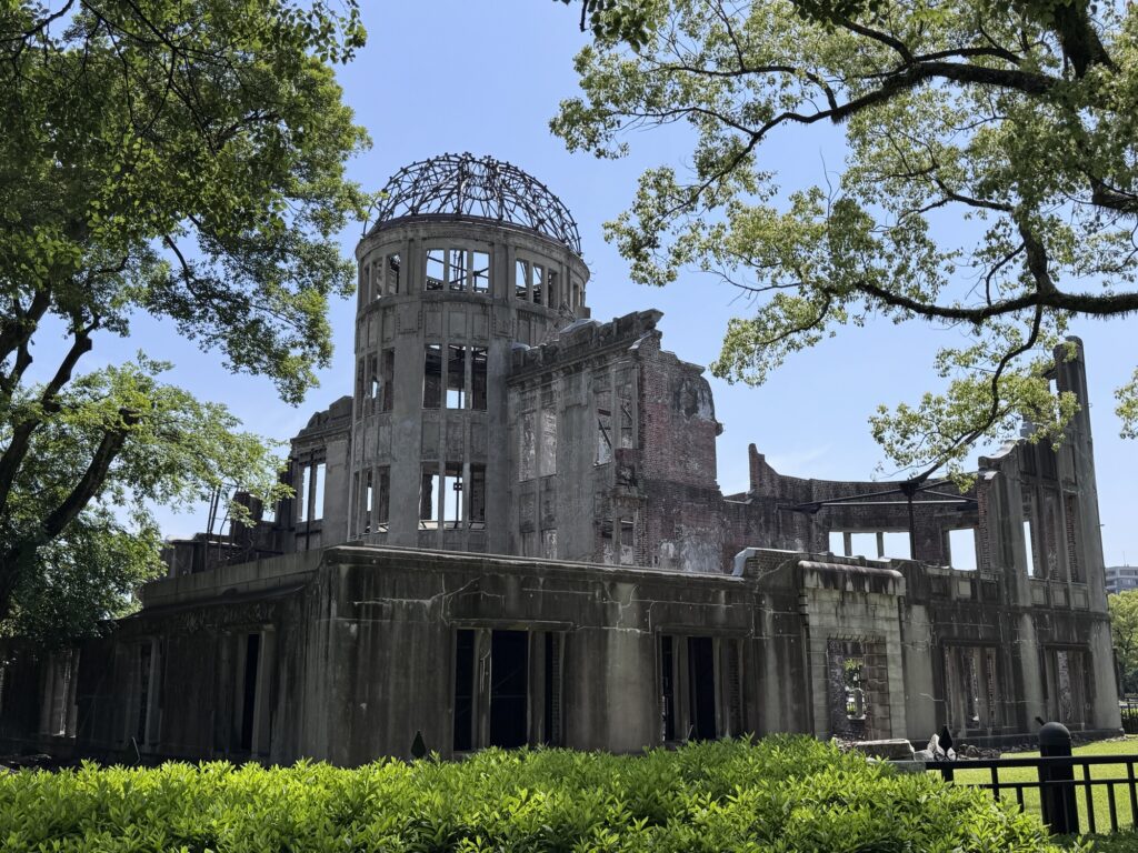 The ruins of the Hiroshima Peace Memorial, also known as the Atomic Bomb Dome, stand under a clear blue sky, surrounded by lush green trees and bushes.