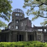 The image shows the ruins of the Hiroshima Peace Memorial, also known as the Atomic Bomb Dome, surrounded by green trees and bushes under a clear blue sky.
