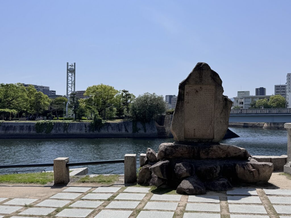A large stone monument stands beside a river under a clear blue sky, with trees, a bridge, and city buildings visible in the background.