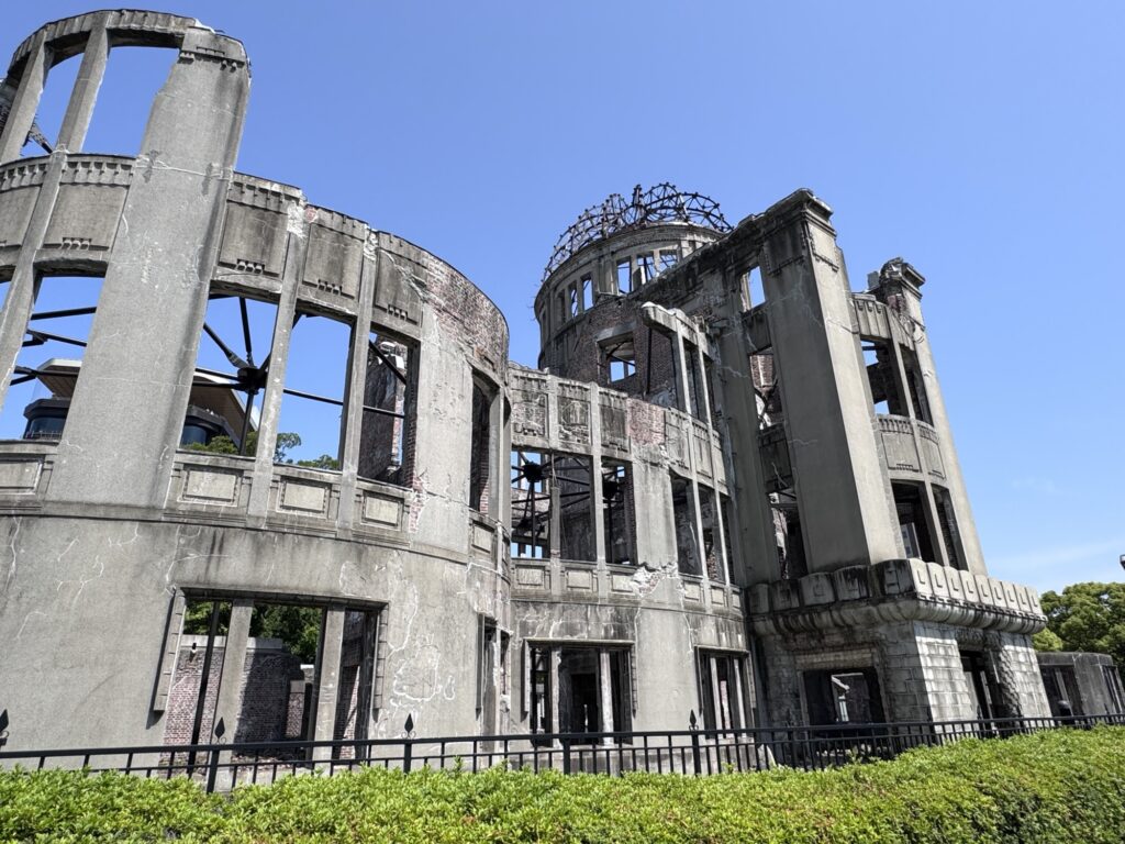 A partially destroyed concrete building with exposed metal dome, surrounded by a low fence and greenery, stands under a clear blue sky. This is the Hiroshima Peace Memorial, also known as the Atomic Bomb Dome.