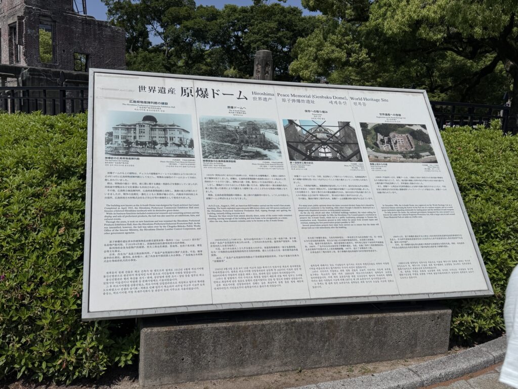 An informational sign about the Hiroshima Peace Memorial (Atomic Bomb Dome) World Heritage Site, featuring text in Japanese and English, historical photos, and detailed descriptions, surrounded by green bushes.