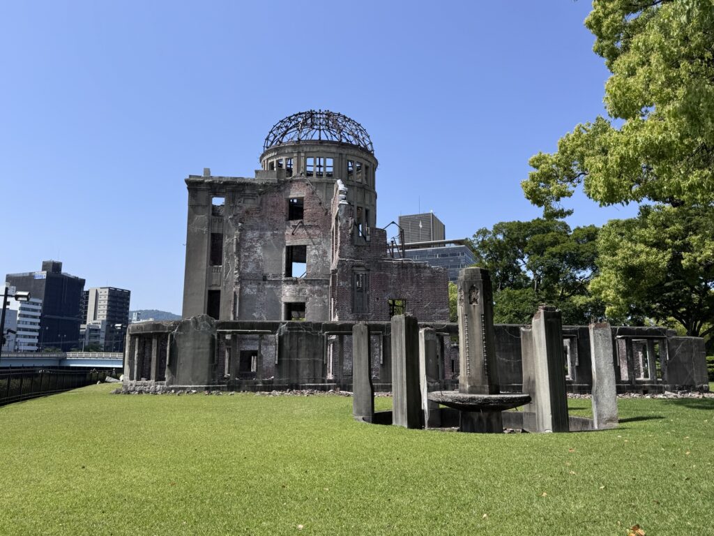 A large, partially destroyed brick building with a domed steel frame on top stands on green grass under a clear blue sky, surrounded by trees and city buildings.