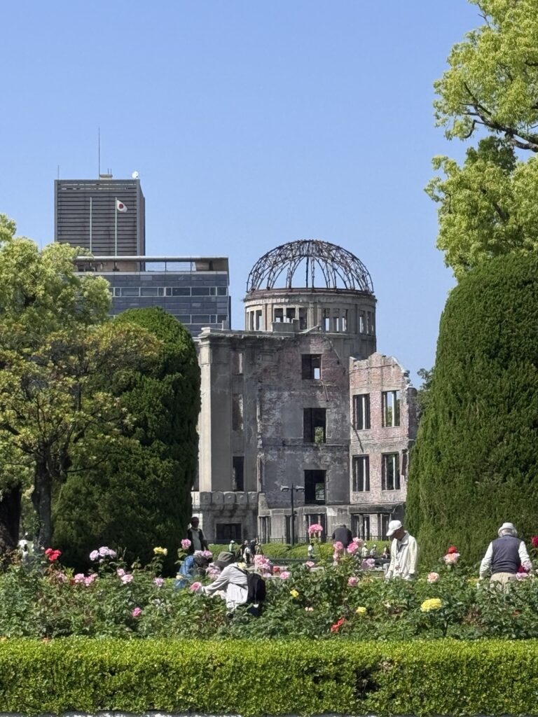 People walk through a garden with blooming flowers and trimmed bushes in front of the ruined Hiroshima Peace Memorial (Atomic Bomb Dome) under a clear blue sky, with modern buildings in the background.