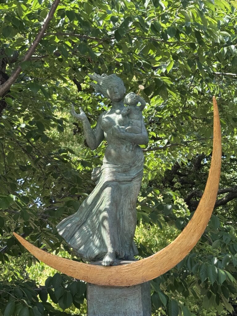 A bronze statue of a woman holding a child stands atop a golden crescent moon, surrounded by lush green trees in the background. The woman’s dress and hair appear to be gently blowing in the wind.