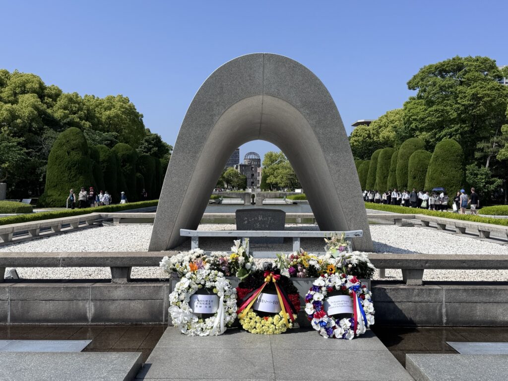 A stone memorial arch at Hiroshima Peace Memorial Park, with flower wreaths placed in front and the Atomic Bomb Dome visible in the background. People stand on walkways surrounded by green trees under a clear sky.