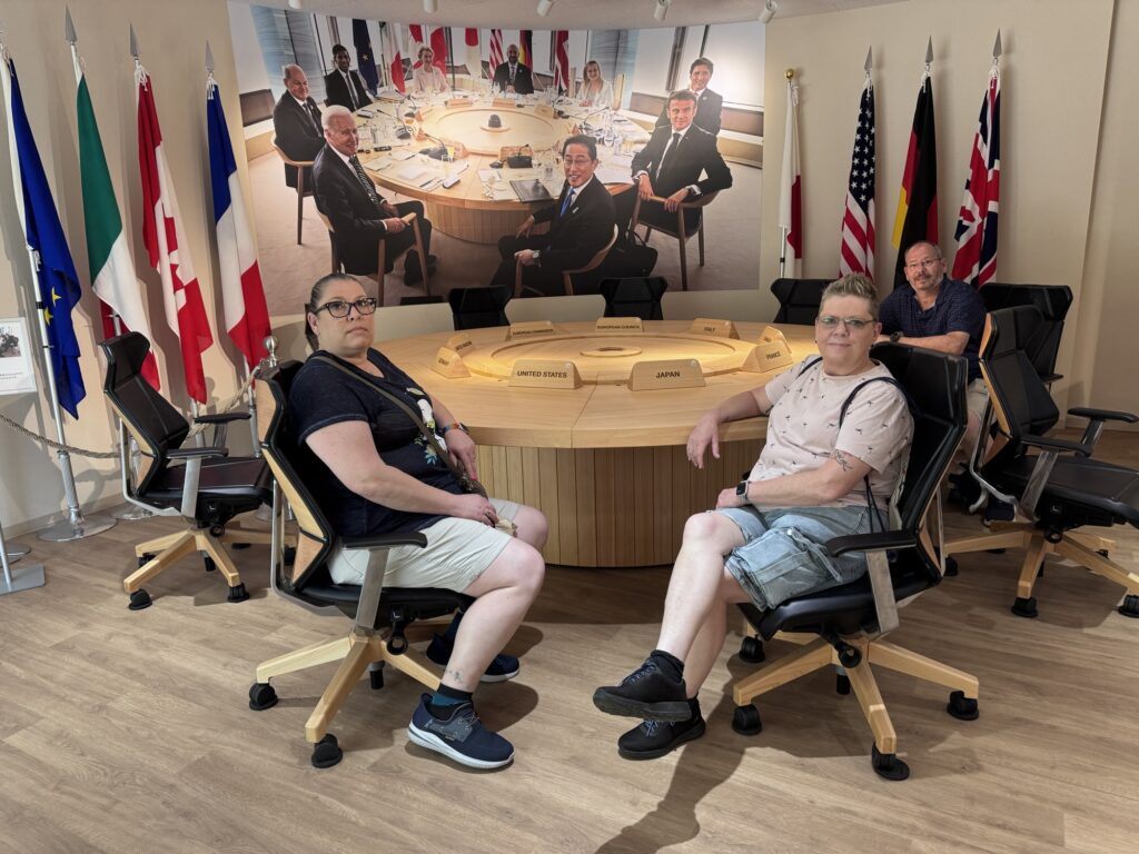 Three people sit around a round table with nameplates and flags in a room. Behind them is a large photo of world leaders at a similar table with more flags, creating a staged, diplomatic atmosphere.