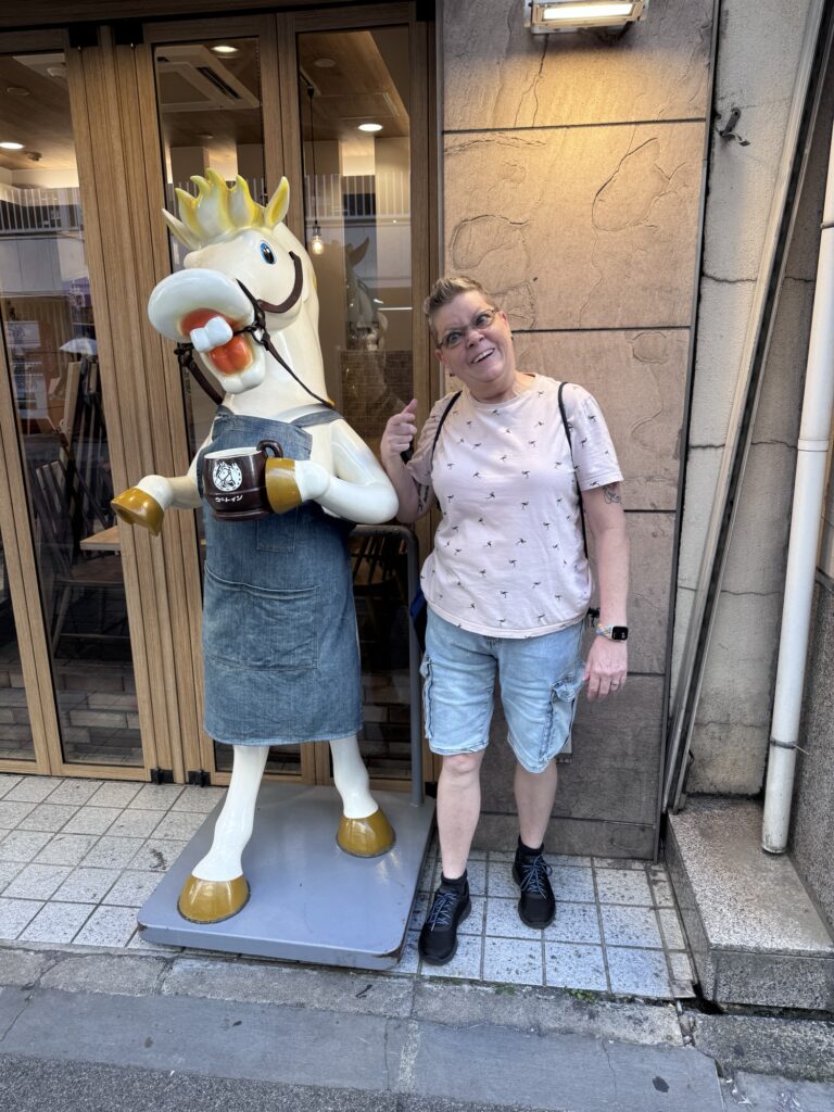A person with short hair and glasses poses and smiles next to a horse statue wearing a crown, apron, and holding a coffee mug, outside a shop with wooden doors and stone walls.