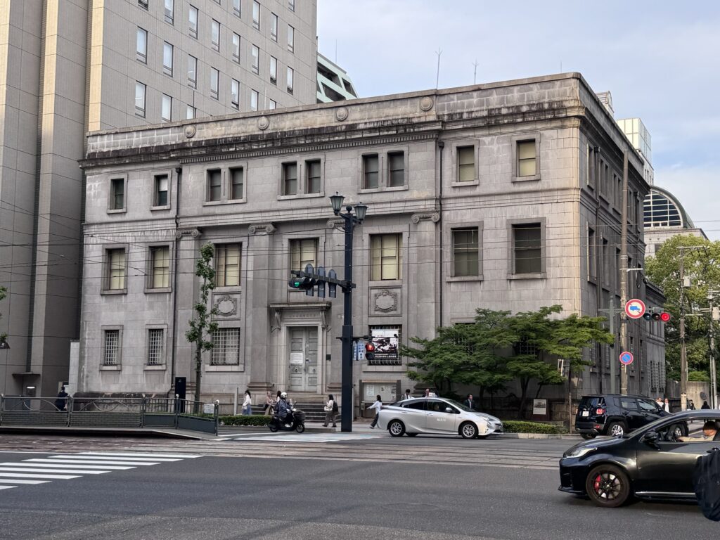 A gray stone building with classical architecture stands at a street corner. Cars, bikes, and people move along the road in front, with modern tall buildings beside and behind it. A tree grows near the entrance.