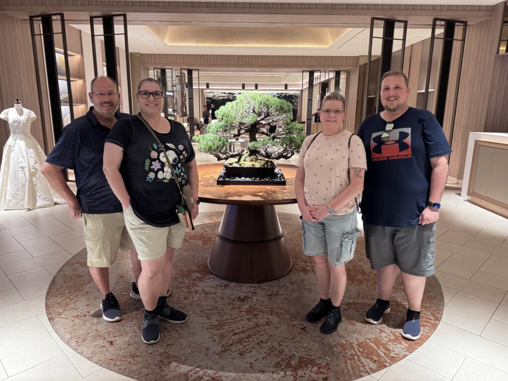 Four adults stand smiling around a circular wooden table displaying a large bonsai tree in an elegant, well-lit room with wood paneling. A mannequin in a white dress is visible on the left side of the image.