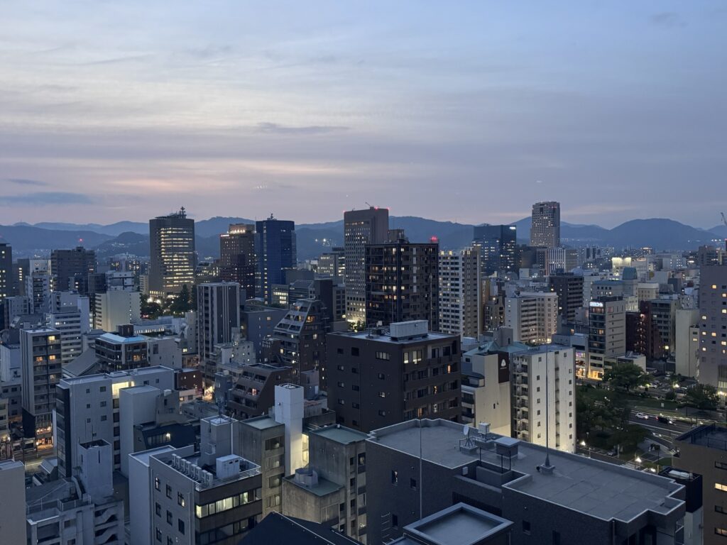 City skyline at dusk with many buildings and apartments; lights starting to turn on. Mountains are visible in the background under a blue and purple sky with scattered clouds.