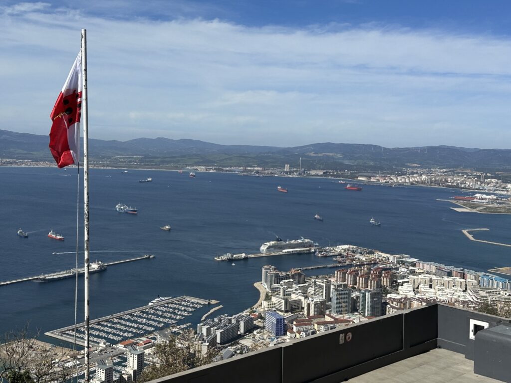 Rock of Gibraltar pathway overlooking the Mediterranean Sea, with visitors walking along a hillside trail and views of ships, coastline, and the mountains of Africa visible in the distance.