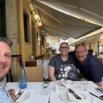 Three people smile at a table set for a meal at an outdoor restaurant. The street is lively, with more diners in the background and buildings painted yellow. Bottled water and glasses are on the table.