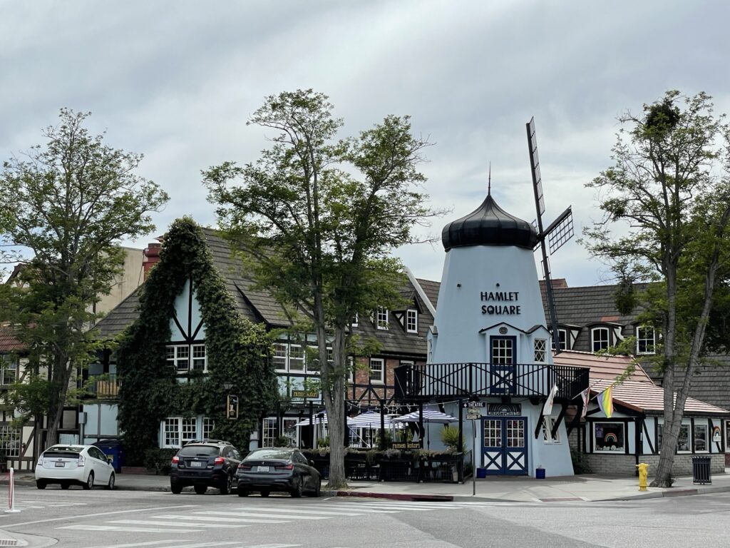 A street view of a quaint European-style building complex with a windmill labeled Hamlet Square, ivy-covered walls, outdoor seating, and parked cars under cloudy skies.