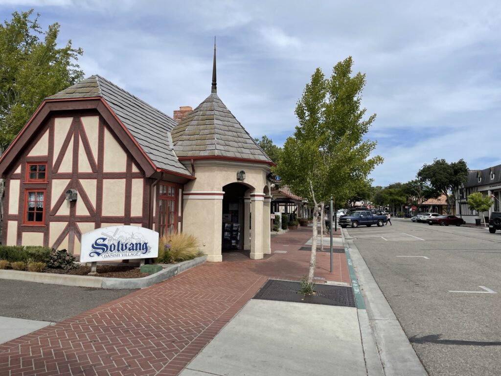 A street view in Solvang, California, shows a building with Danish-style architecture, a sign reading Solvang, brick sidewalks, trees, and parked cars under a partly cloudy sky.