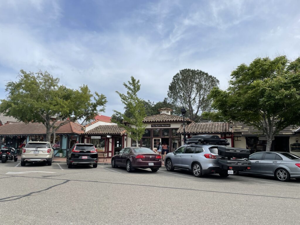 A row of parked cars lines the street in front of small shops with red-tiled roofs and large windows, set among green trees under a partly cloudy sky.