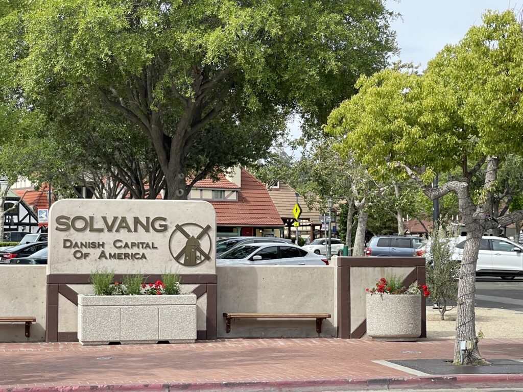 A sign reading Solvang Danish Capital of America with a windmill icon stands in front of trees, benches, and buildings with red-tiled roofs in a quaint downtown area.