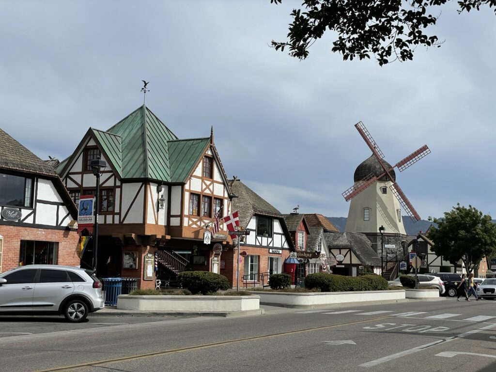 A street scene in Solvang, California features Danish-style buildings, a windmill, Danish flags, parked cars, and trees under a partly cloudy sky.