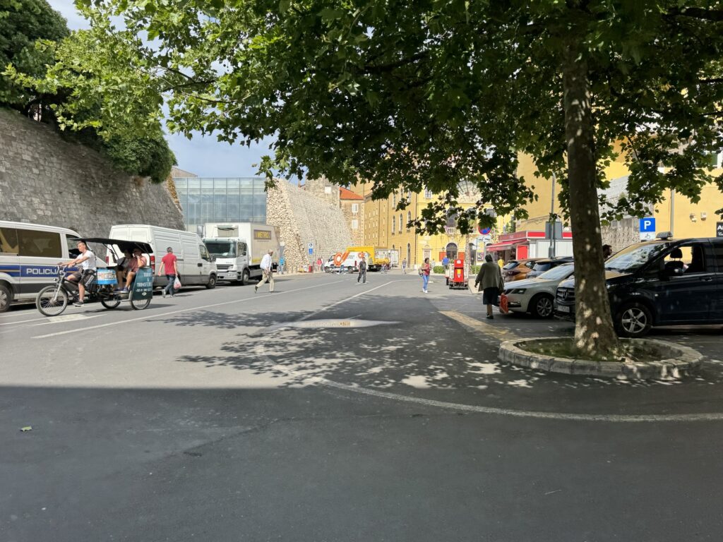 People walk and cycle along a sunny street lined with parked cars and vans. A leafy tree provides shade in the foreground, and stone walls and yellow buildings are visible in the background.