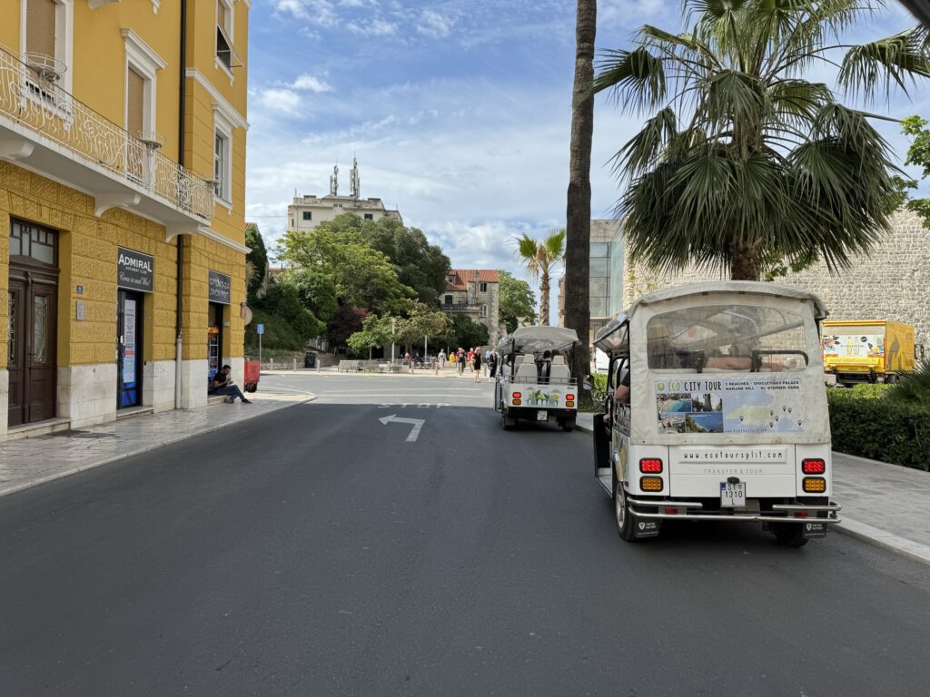 Two small open tour vehicles drive down a paved street lined with yellow buildings, palm trees, and greenery under a partly cloudy sky. People are visible in the background near a park area.