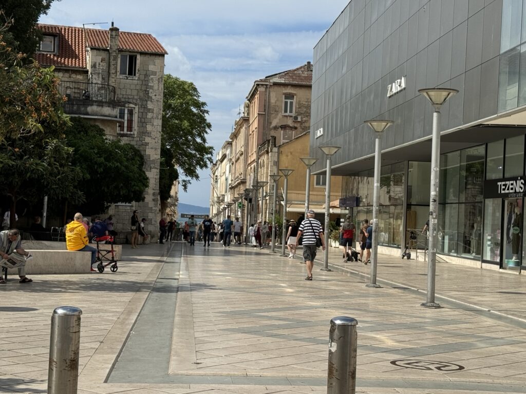 A pedestrian street with people walking and sitting on benches, flanked by modern shops on the right and older buildings on the left, with trees and a partly cloudy sky in the background.
