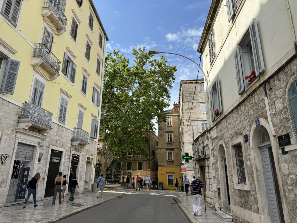 A narrow street in a European city is lined with stone buildings, shops, and balconies. People walk along the sidewalk. A large tree and a green pedestrian light are visible under a partly cloudy sky.