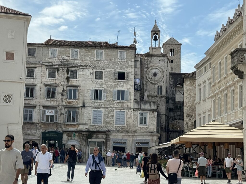 People walk through a sunny plaza surrounded by historic stone buildings, including one with a large clock and bell tower. Outdoor café tables with umbrellas are on the right, and the sky is partly cloudy.