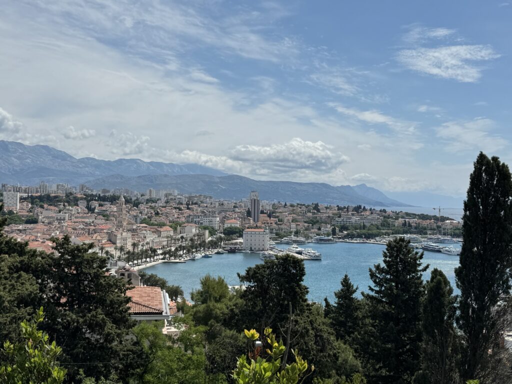 View of a coastal city with red-tiled roofs, a harbor with yachts, and mountains in the background, under a partly cloudy sky, framed by trees and greenery in the foreground.
