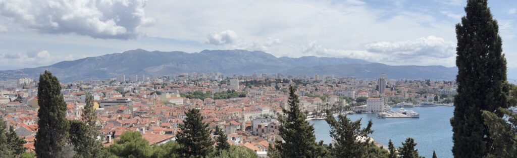 Panoramic view of a coastal city with red-roofed buildings, a marina with boats, and mountains in the background under a partly cloudy sky, framed by tall evergreen trees.